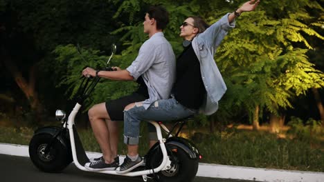 A-happy-couple-riding-on-electro-bike-in-street-on-summer-day.-Beautiful-girl-and-her-boyfriend-enjoying-their-time.-Park-territory