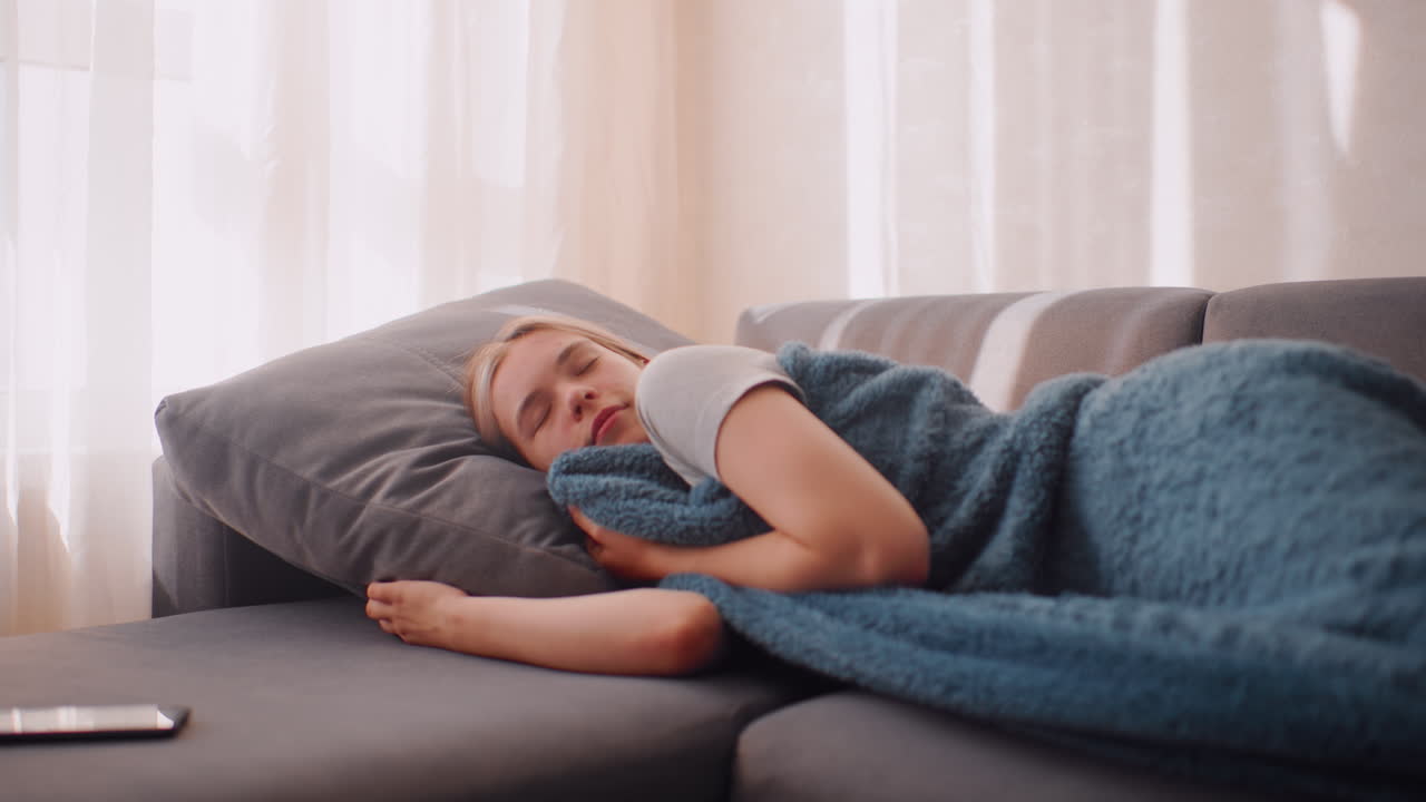 Young lady holding blanket warmly while sleeping on couch resting head on pillow enjoying peaceful comfort as sunlight streams through curtains creating serene indoor atmosphere