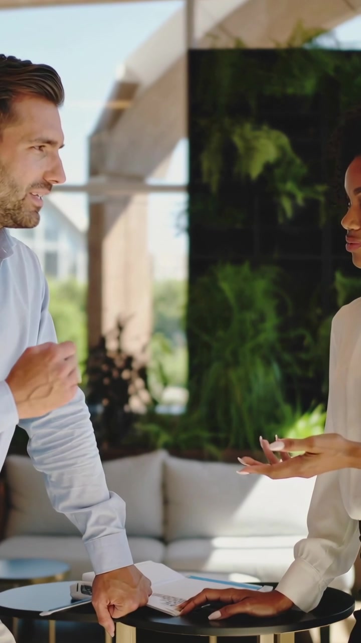 Vertical video: Pointing man in blue shirt and woman in white blouse reviewing documents in lounge