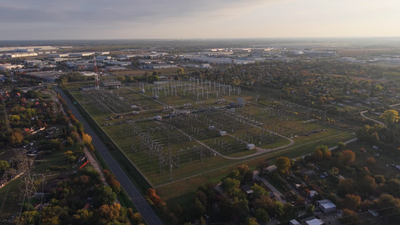An aerial video of an electrical substation surrounded by industrial buildings and residential areas. The video is taken during sunset, highlighting the power infrastructure and transmission lines