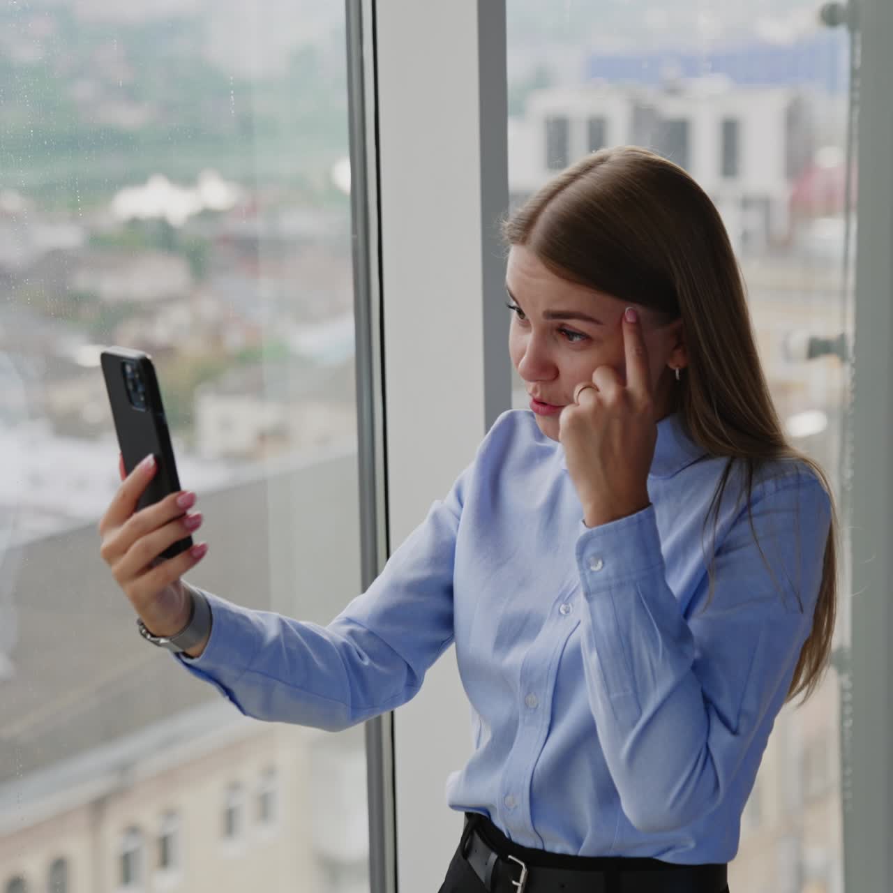 Good-looking blonde lady wearing official clothes has video-call. Woman stands near a big window communicating via video chat