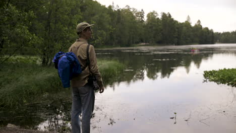 Man-with-backpack-and-camera-looks-out-on-lake-and-forest,-slow-pan