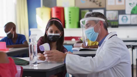 Diverse-male-teacher-showing-schoolgirl-how-to-measure-temperature,-all-wearing-face-masks