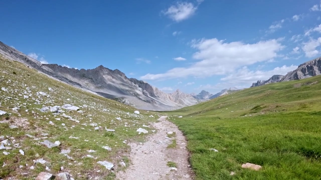 Un sentiero sterrato roccioso si snoda attraverso un'ampia valle alpina d'alta quota sotto un cielo estivo luminoso, con cime aspre in lontananza