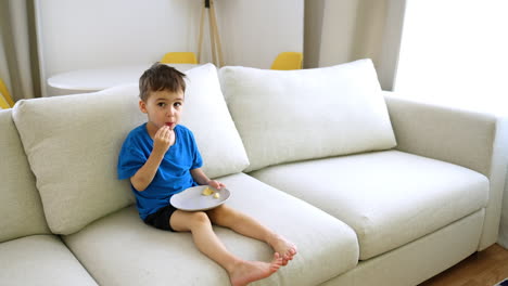 Child enjoying snacks on a couch. A young boy sits comfortably on a couch, happily eating snacks while relaxing in a well-lit living room