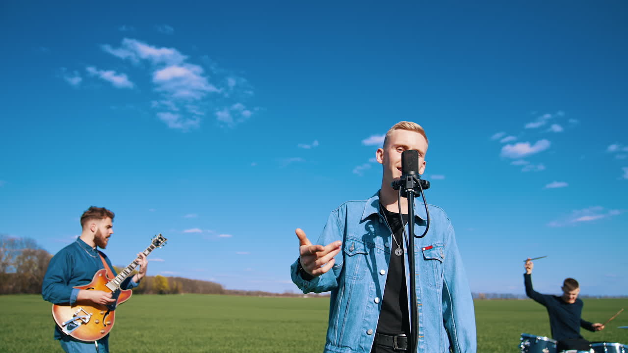 Young music group performing on blue sky background. Male singer singing into a microphone while musical band playing instruments on the field.