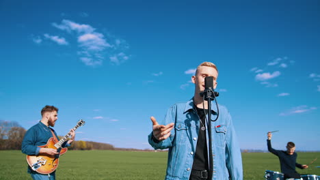 young music group performing on blue sky background. male singer singing into a microphone while musical band playing instruments on the field.