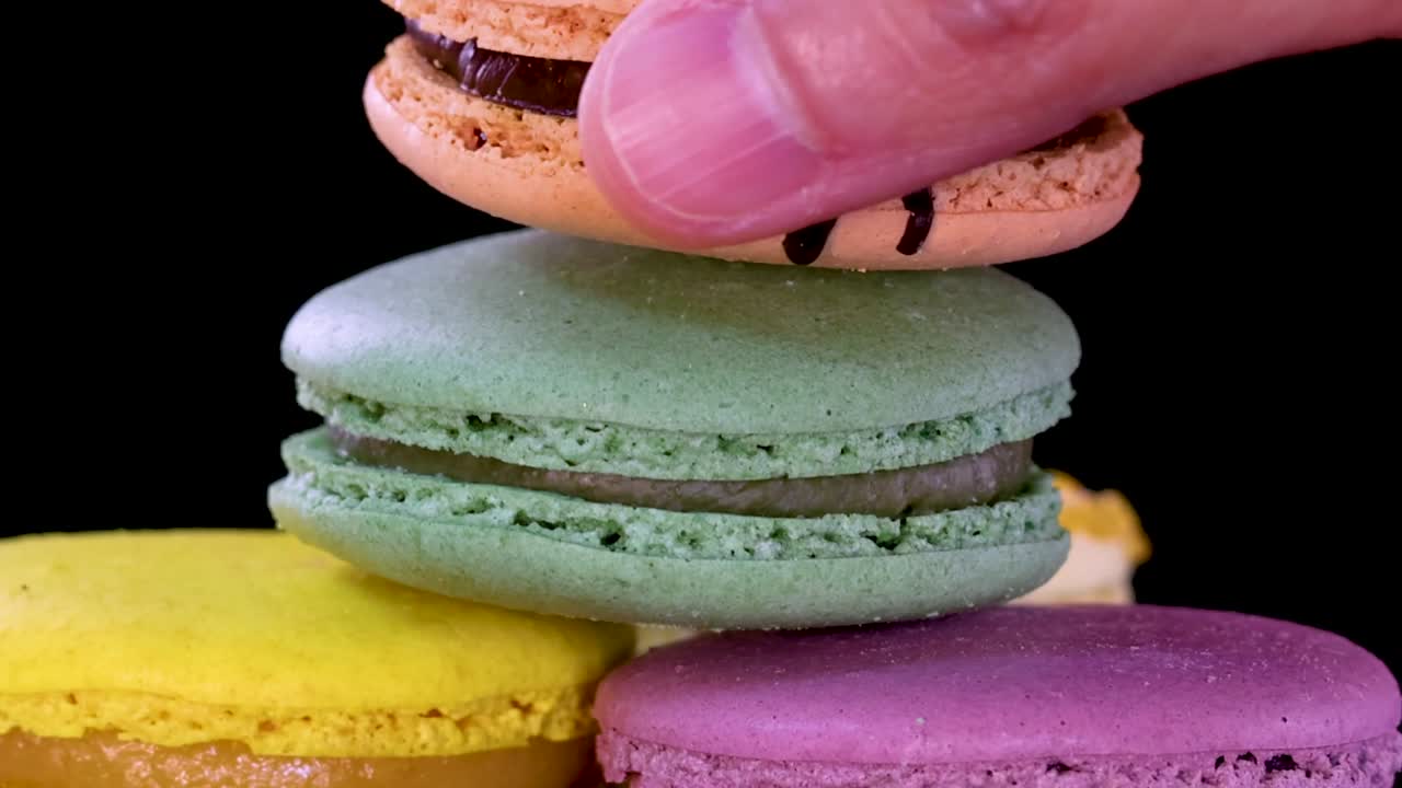 A hand carefully stacks vibrant green, yellow, and purple macarons against a dark background