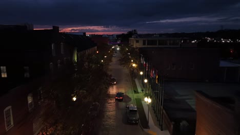 night scene in american town driving car on downtown road at night. warm lights of lantern along street. rising drone tracking shot. historic and ancient buildings in center of city. wide shot.