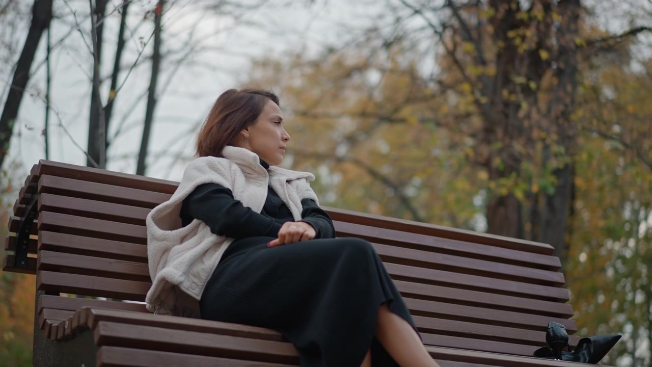 Giovane donna si appoggia all'indietro su una panchina di legno del parco con le gambe incrociate e le mani in grembo, guardando a sinistra con un'espressione calma e riflessiva nella soffusa luce autunnale nuvolosa all'aperto, circondata da alberi spogli.