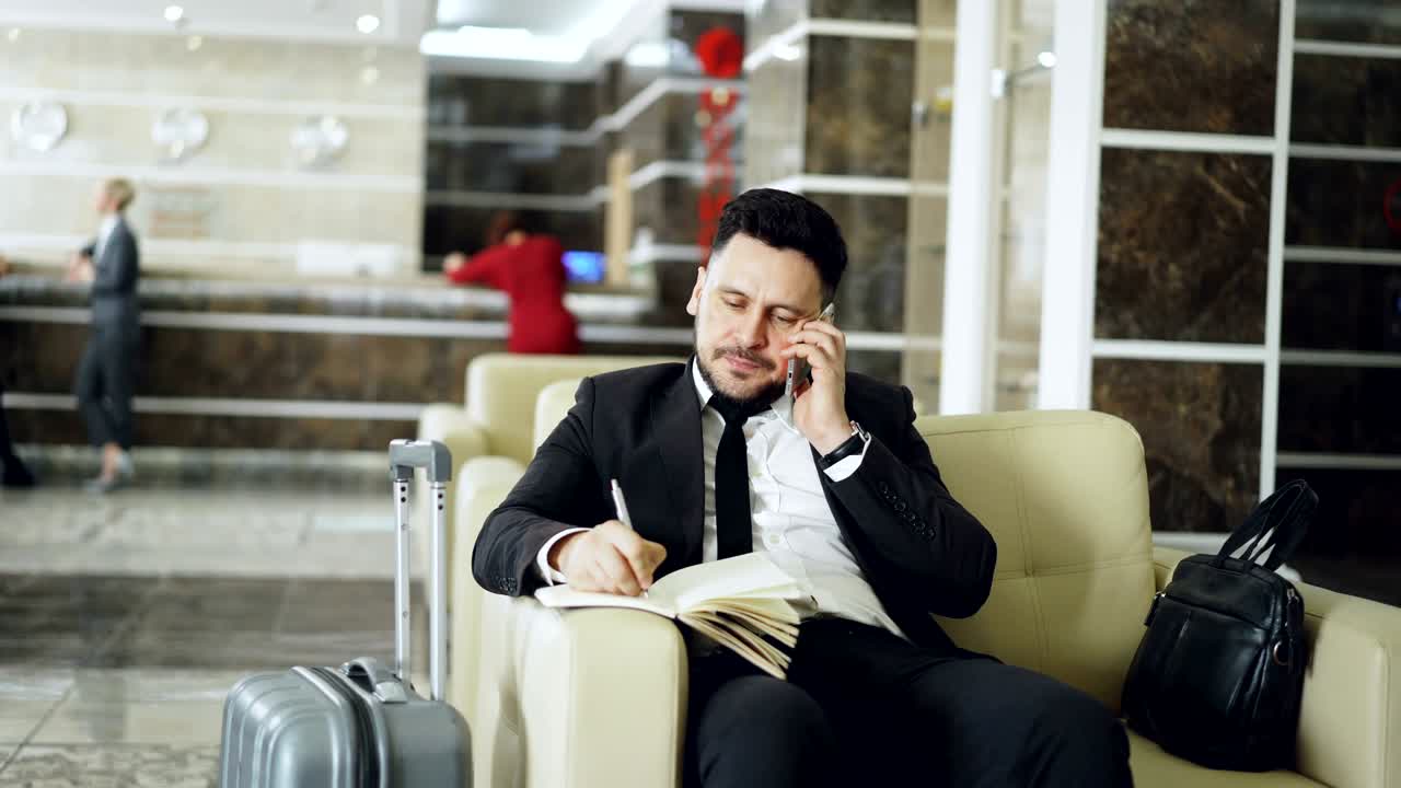 tomada de un hombre de negocios confiado hablando por teléfono móvil y escribiendo notas en el bloc de notas mientras está sentado en un sillón en un hotel de lujo con equipaje cerca de él. concepto de viaje, negocios y personas