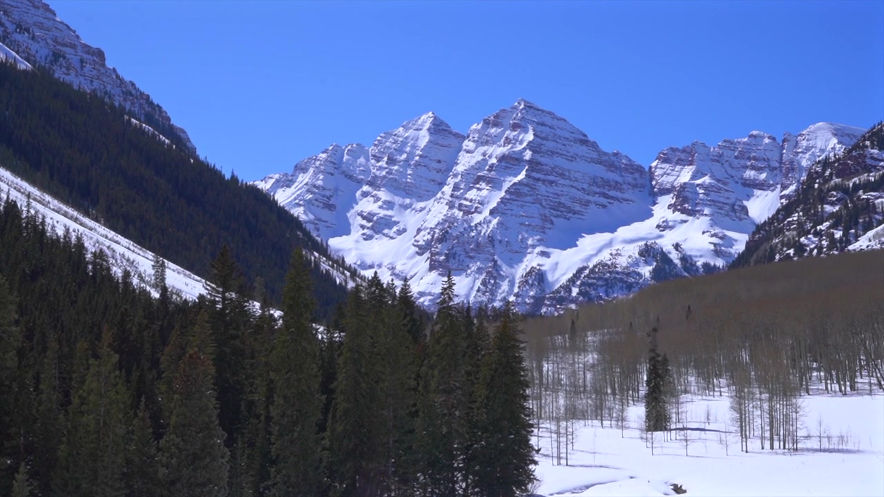 Vista mattutina con teleobiettivo delle cime delle Montagne Rocciose Maroon Bells (14er) sopra una valle innevata e una fitta foresta di pioppi tremuli sempreverdi, che cattura la nitida luce mattutina invernale, un classico paesaggio montano del Colorado