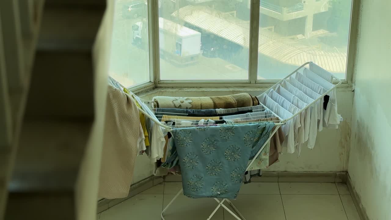 Clothes hang on a folding drying rack beside large windows in daylight, showing an indoor laundry area with everyday garments, tiled floor, and a practical domestic setting