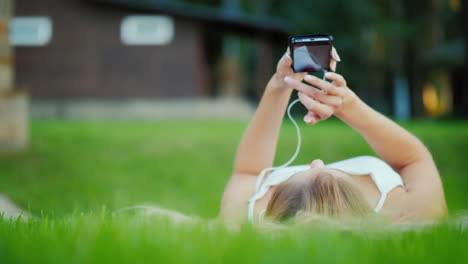 A-Young-Girl-Is-Lying-On-Her-Back-On-The-Lawn-Enjoying-A-Smartphone-Video-With-Shallow-Depth-Of-Fiel
