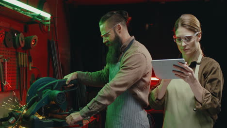 joven soldadora caucásica con gafas y delantal tocando y desplazándose en un dispositivo de tableta mientras observa a su compañero de trabajo cortando metal en la máquina en el taller