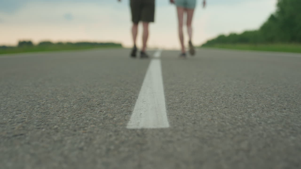blurred lower bodies of man and woman walking hand in hand along white road centerline flanked by green fields under late afternoon sky conveying motion connection adventure outdoors