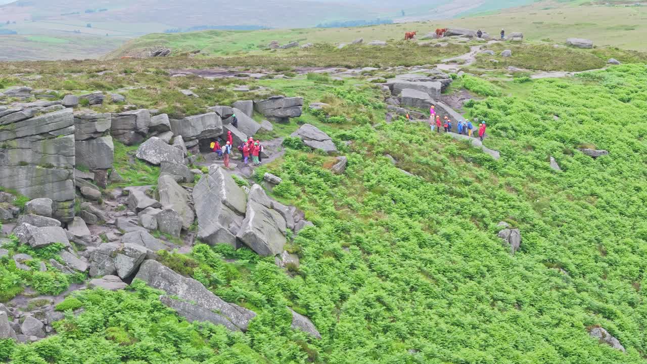 A group of climbers in red jackets explores a rocky moorland under an overcast sky. The scene captures the essence of outdoor adventure, with rugged terrain and expansive views