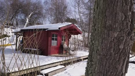 Zoom-in-aerial-shot-of-rustic-cabin-next-to-frozen-lake-with-young-woman-enjoying-the-views-from-the-deck