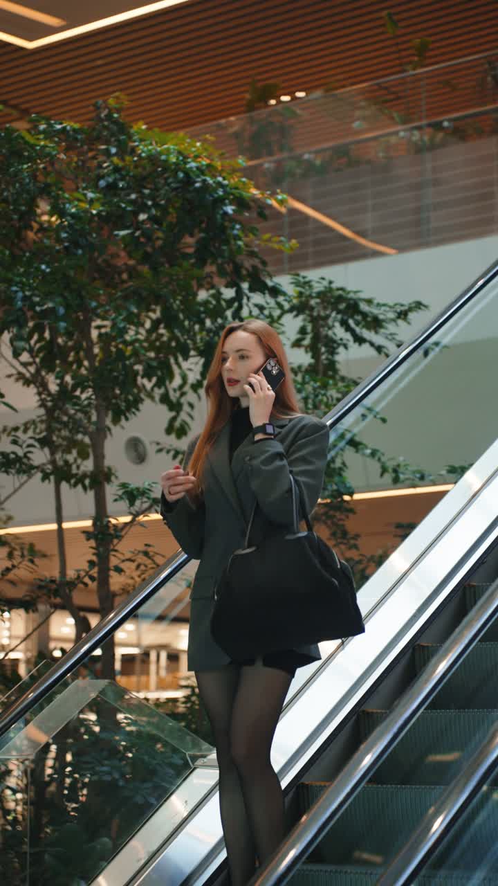 A successful young businesswoman in a stylish blazer talks on her cell phone while riding an escalator in a contemporary building, representing a busy, on-the-go corporate lifestyle.