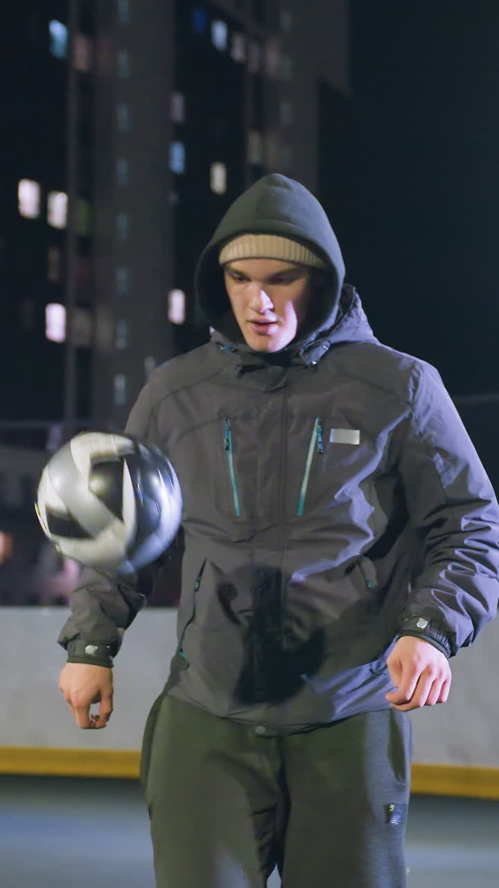 Coach in hoodie joggling football with focus and precision during nighttime training on outdoor field, illuminated by urban lights and surrounded by high-rise buildings and fences