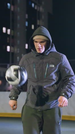 entrenador con capucha joggling fútbol con enfoque y precisión durante el entrenamiento nocturno en el campo al aire libre, iluminado por luces urbanas y rodeado de edificios de gran altura y cercas