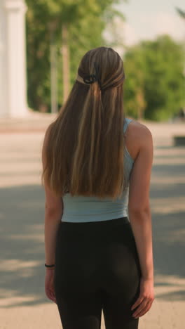 vista trasera de una joven con pantalones cortos negros y camiseta cian caminando tranquilamente por un camino entrelazado con un fondo escénico con edificios históricos, bancos, postes de lámparas, vegetación exuberante bajo la cálida luz del sol