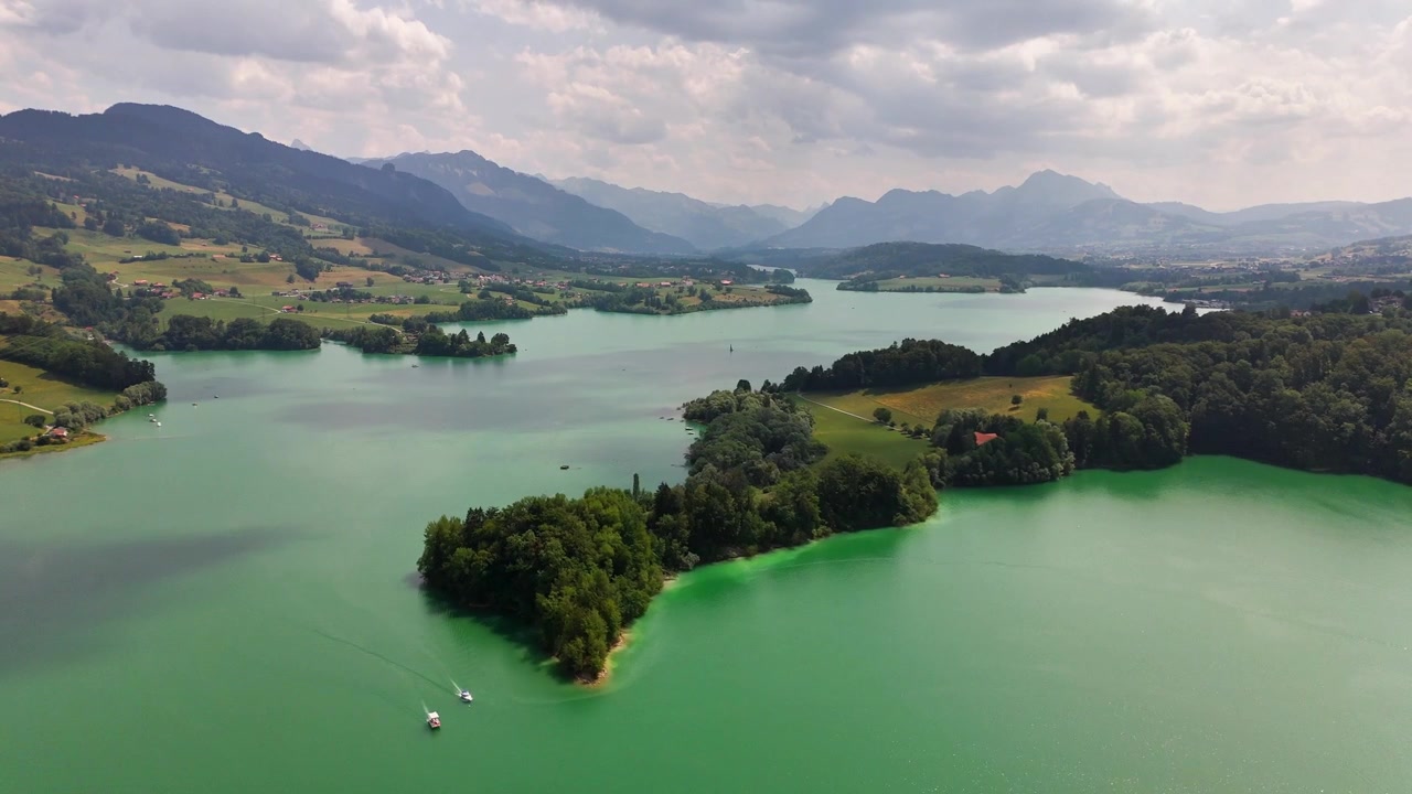 Der Greyerzersee nahe Pont-la-Ville, Freiburg, Schweiz, zeigt bewaldete Inseln, ein türkisfarbenes Reservoir, sanft gewelltes Ackerland und dahinter die Freiburger Voralpen, langsames Herauszoomen der Drohne.
