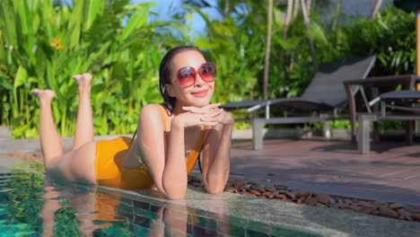 Attractive-hot-Asian-Woman-Lying-on-her-Belly-on-the-Border-of-the-Swimming-Pool-of-Tropical-Hotel-Looking-Straight-Into-the-Camera-Lens-Smiling-While-Moving-Legs-Up-and-Down,-slow-motion