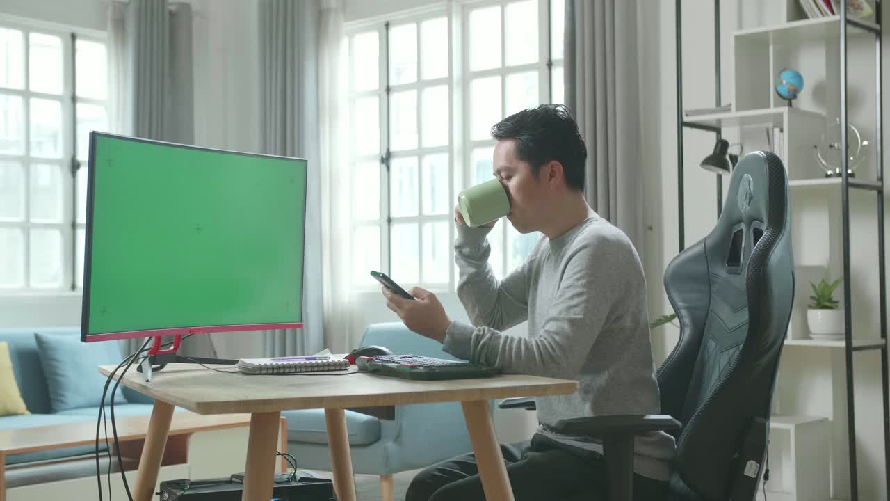 Asian Man In Long Sleeved T-Shirt And Black Pants With Green Screen Desktop Computer Using Mobile Phone And Drinking Coffee While  Working At Home.