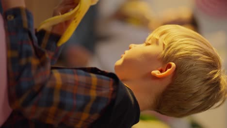 Vertical-video-shooting-close-up-of-a-stole-boy-with-blue-hair-eating-a-banana-during-a-break-and-lunch-in-a-club-for-preschool-children-in-a-sunny-cozy-room