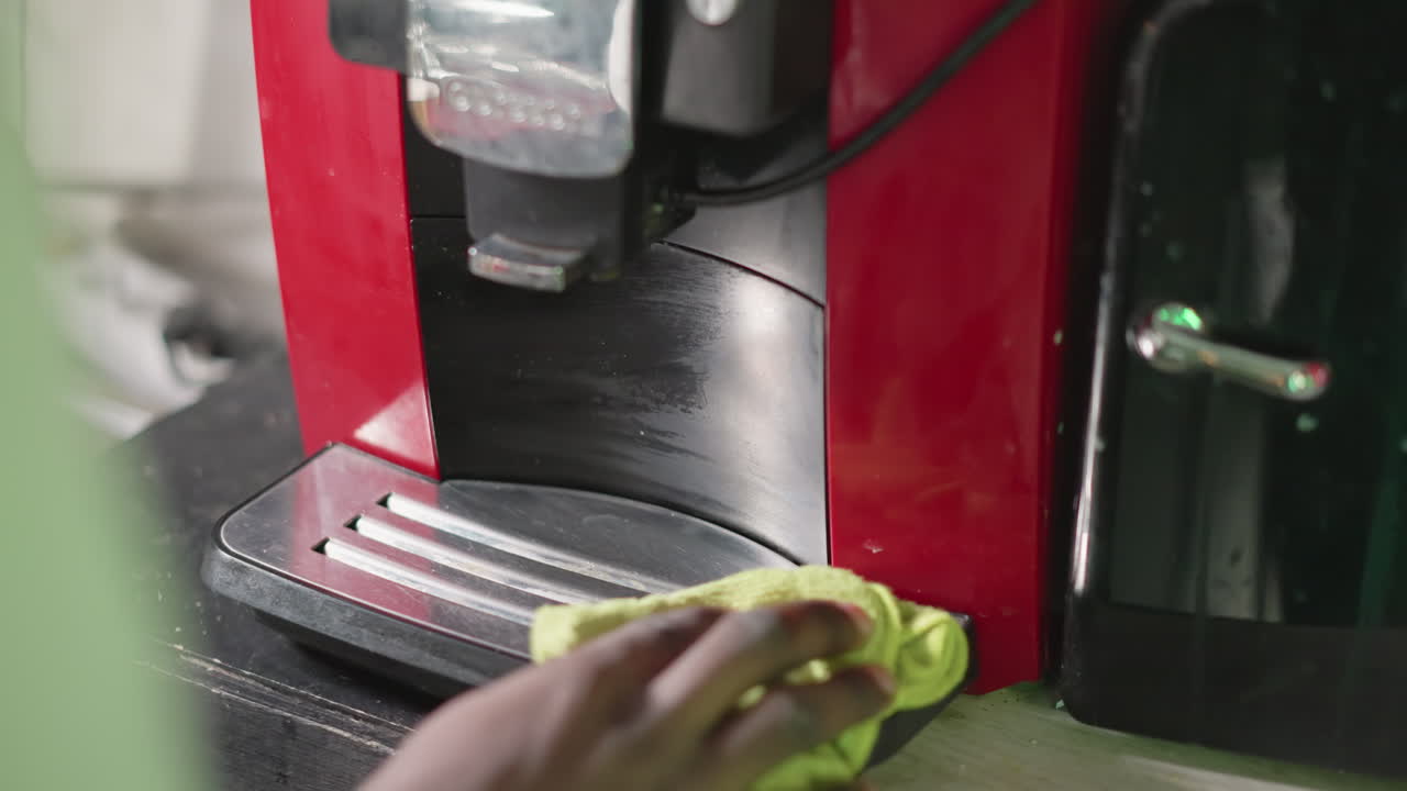 Close-up of hand cleaning red coffee machine with yellow cloth, wiping down front surface of appliance, focusing on maintaining cleanliness and appliance