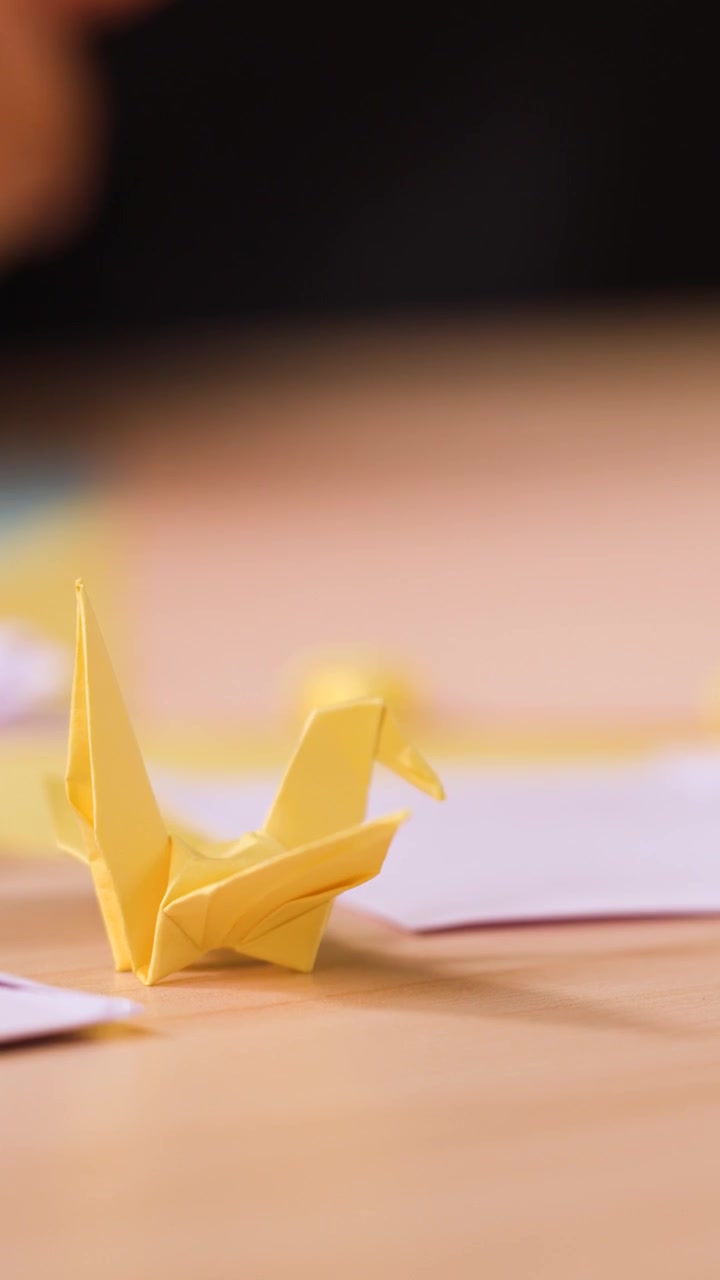 A yellow paper crane sits on a wooden desk with blurred hands crafting in background