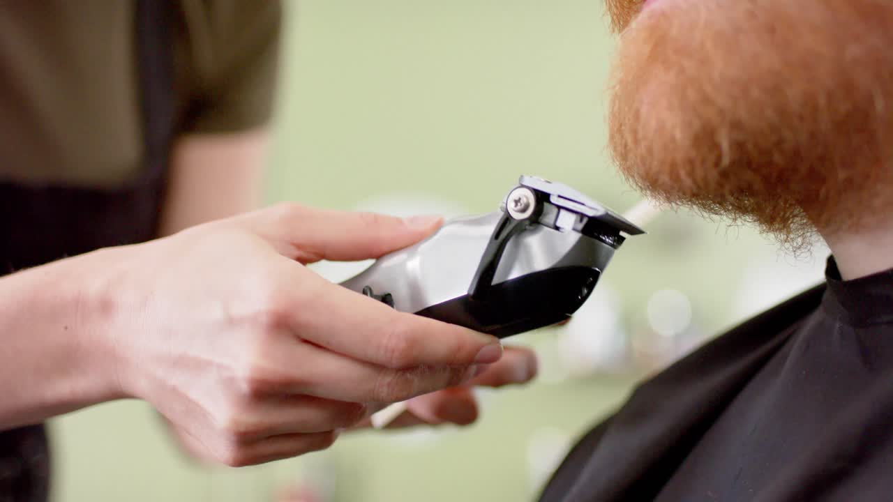Hænderne på en kaukasisk kvindelig barber, der trimmer en mandlig kundes røde skæg i barbersalonen, i slowmotion.
