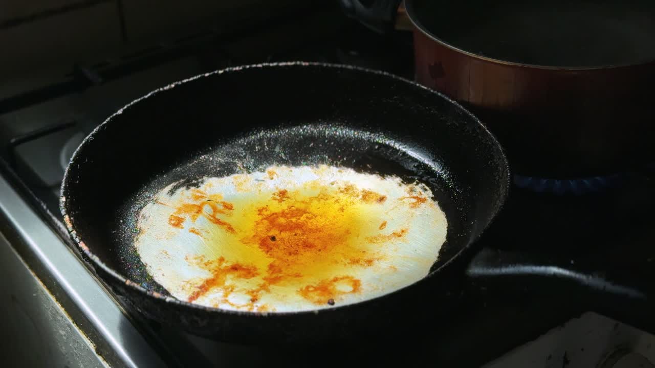 Close-up minyak goreng panas yang sedang memanas di wajan hitam di atas kompor gas, dengan panci logam di latar belakang, memperlihatkan penataan dapur dalam ruangan untuk persiapan dan memasak makanan.