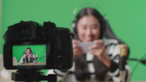 close up of a camera monitor recording asian woman gamer with headphone and computer using mobile phone playing game on the green screen background