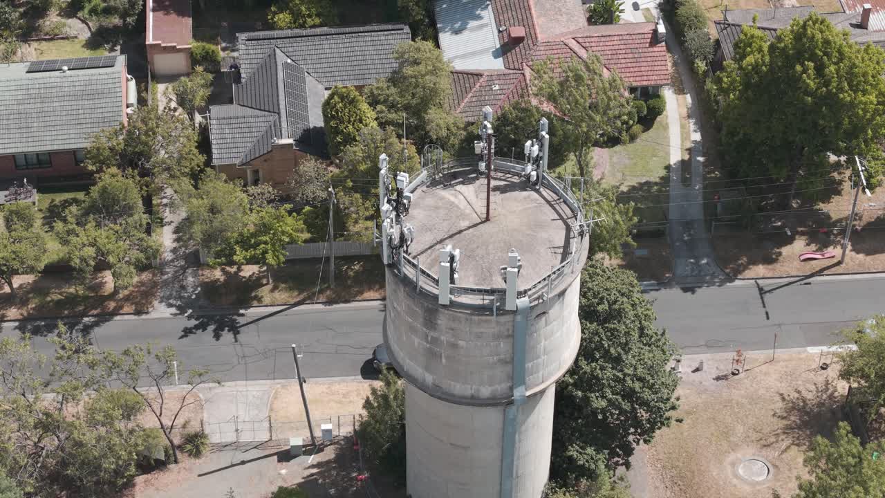 Drohnenaufnahme, die einen Wasserturm mit 5G-Antennen zeigt, umgeben von einer üppigen Vorstadtlandschaft. Die Luftperspektive betont die technologische Integration im Alltag.