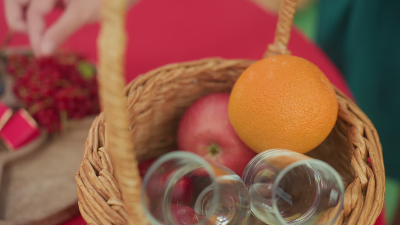 mani che dispongono mele arancioni in un cesto, cesto di vimini posto su una tovaglia rossa con bicchieri, una ciotola di albicocche e ribes nelle vicinanze, mani delicate che selezionano la frutta per un intimo picnic, atmosfera calda