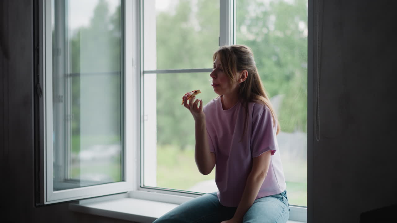 Lady seated on window ledge savoring pizza slice, wearing casual purple t-shirt and jeans, enjoying peaceful moment near open window with blurred greenery background on bright day