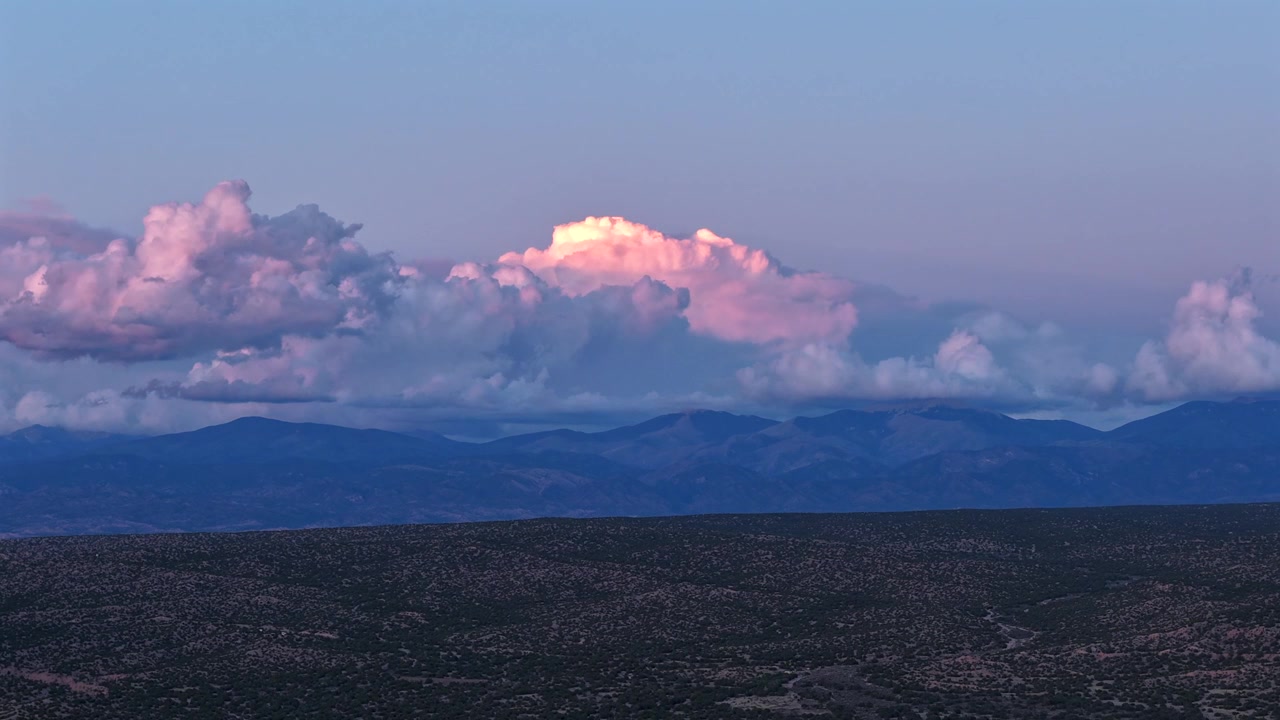 Sää-timelapse tallentaa erottuvan valkokärkisen pilven liikkuvan täyteläisten purppuranväristen auringonlaskun värikerrosten yläpuolella. Muutos tapahtuu karujen vuorten siluetin yllä lounaassa.