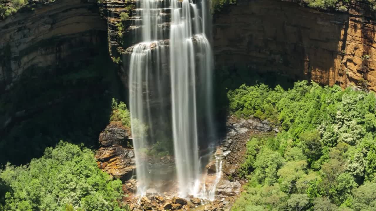 Filmagem em time-lapse de uma cachoeira alta caindo por um penhasco íngreme em uma área rochosa cercada por densa vegetação verde. Múltiplos cursos de água fluem rapidamente