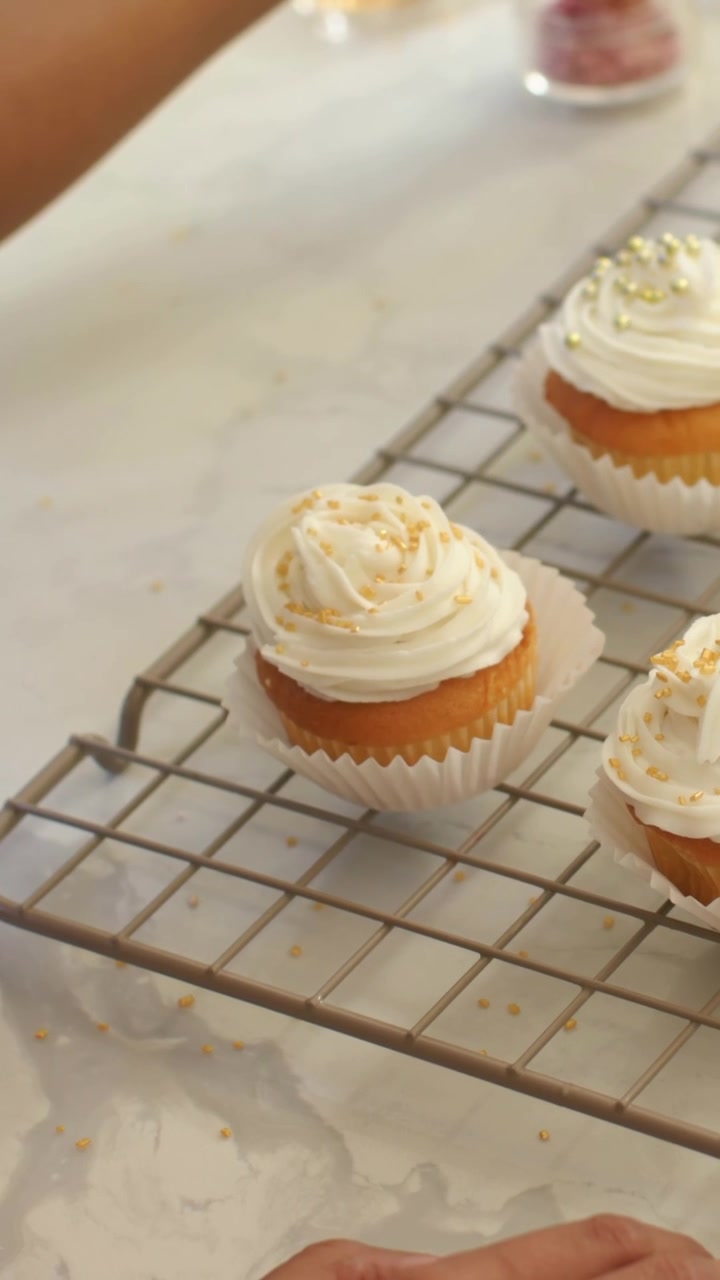 Video verticale: Una mano entra nell'inquadratura e finisce di decorare un cupcake su una griglia di raffreddamento, mentre la persona indossa un top rosa.