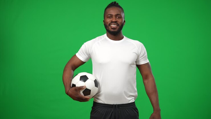 un sportif afro-américain souriant avec un ballon de football marchant de l'arrière-plan à chromakey mockup regardant autour et regardant la caméra. portrait d'un homme confiant positif posant sur l'écran vert.