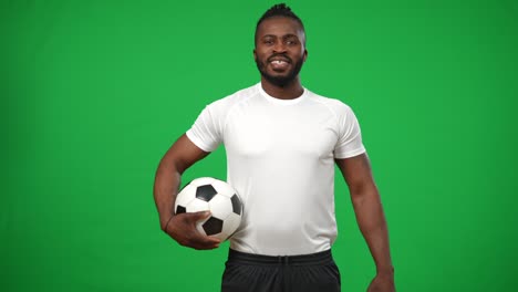 un deportista afroamericano sonriente con una pelota de fútbol caminando desde el fondo en una maqueta de chromakey mirando a su alrededor y mirando a la cámara. retrato de un hombre confiado y positivo posando en la pantalla verde.