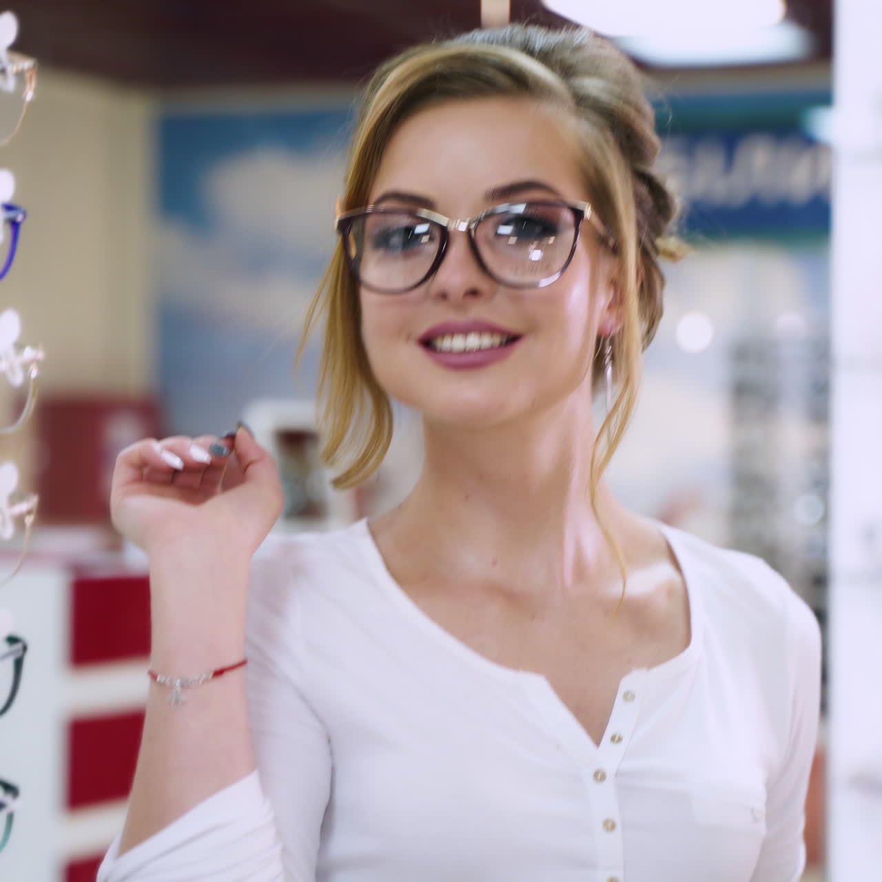 Pretty woman in new glasses poses in front of the mirror. Young model wearing stylish eyeglasses at optical store and smiles happily.