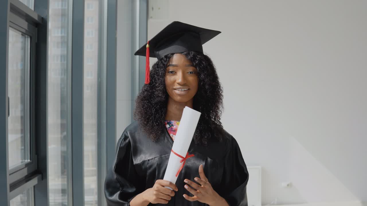 joven graduada afroamericana de pie frente a la cámara con un diploma en las manos. la estudiante lleva una túnica negra y un sombrero de maestro cuadrado.