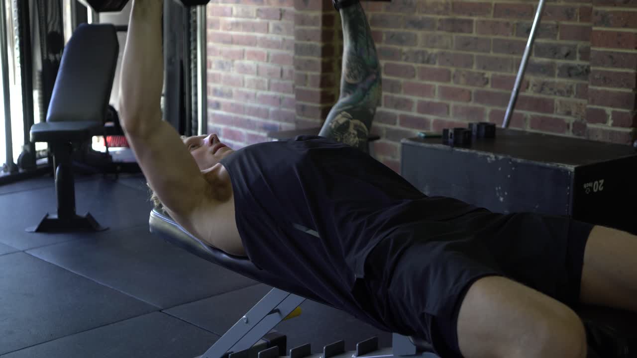 Tattooed man in home gym shot from side doing dumbbell chest press