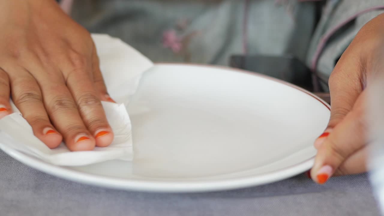 la mano de una mujer con esmalte de uñas naranja limpiando un plato blanco con una servilleta blanca