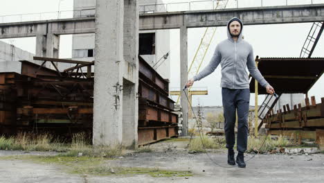 fernsicht auf einen kaukasischen sportler mit bart in einem grauen hoodie, der an einem bewölkten morgen im freien eine verlassene fabrik seilspringt