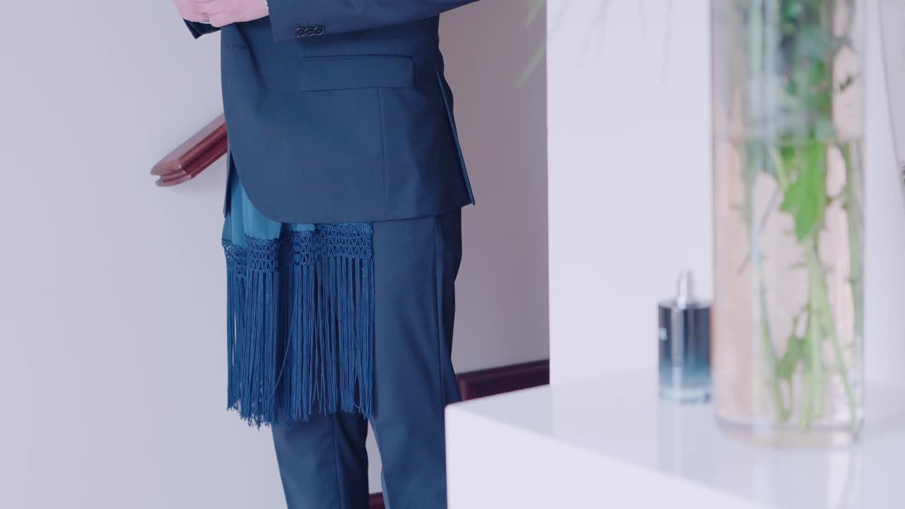 close up of elegant blue suit featuring a long fringed scarf draped at the waist indoors