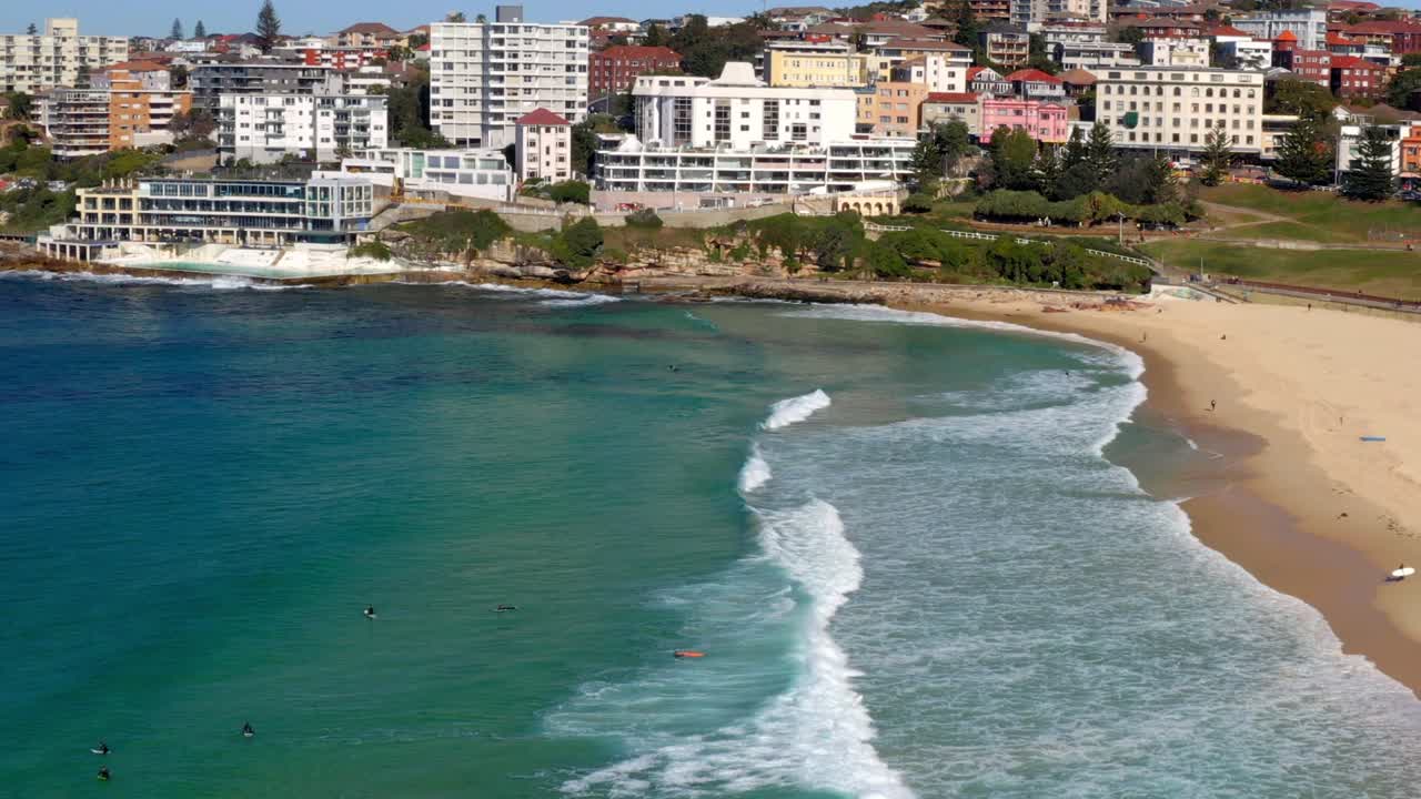 bañistas nadando y surfeando en bondi beach con piscina junto al mar y edificios en segundo plano